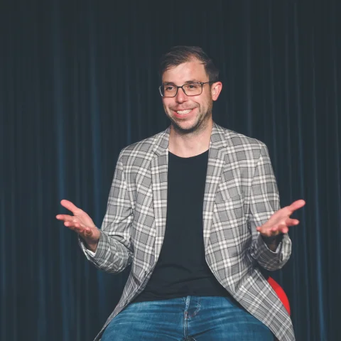 Christian Wirth lacht und gestikuliert mit einem Mikrofon vor einem blauen Bühnenvorhang.